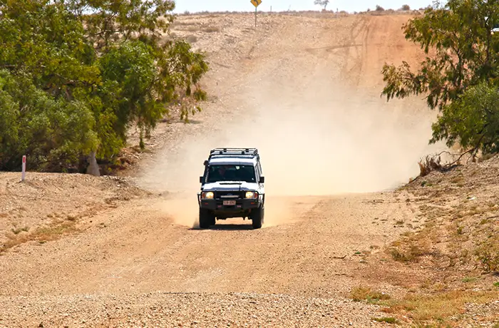 White 4WD vehicle driving on a dusty gravel road in a rural area, kicking up a cloud of dust as it travels between scattered trees.